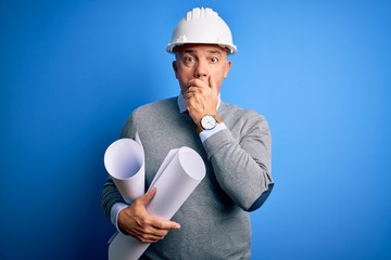 Middle age handsome grey-haired architect man wearing safety helmet holding blueprints shocked...