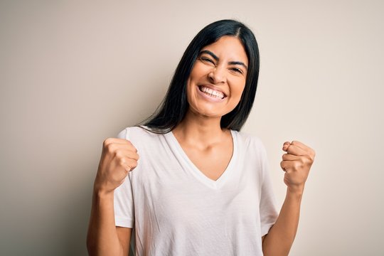 Young beautiful hispanic woman wearing casual white t-shirt over isolated background very happy and excited doing winner gesture with arms raised, smiling and screaming for success. Celebration