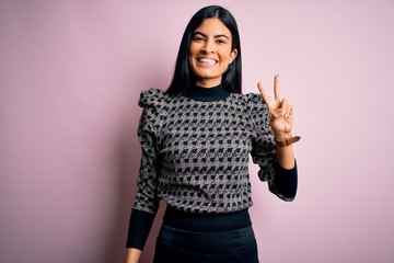 Young beautiful hispanic business woman wearing elegant sweater over pink background smiling with happy face winking at the camera doing victory sign. Number two.