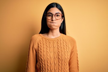 Young beautiful hispanic woman wearing glasses over yellow isolated background puffing cheeks with funny face. Mouth inflated with air, crazy expression.