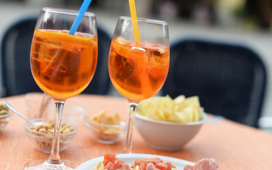 Two bright orange cocktails with ice cubes and colorful straws on a table with crisps and other finger food. Selective focus