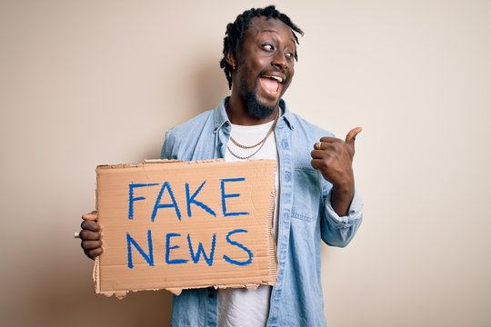 Young african american man holding banner with fake news message over white background pointing and showing with thumb up to the side with happy face smiling