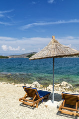 Rocky beach on the shore of the Ionian Sea with sunbeds and straw umbrellas. Blue and tourquse sea in background, nobody. Sunny Summer day. Ksamil, Albania, Europe.