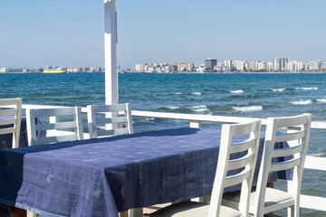 Fototapeta premium Restaurant on the pier in Vlore, Albania on the Ionian Sea, tables covered with blue tablecloths, white chairs on a sunny summer day. Vlora coast line in the background.
