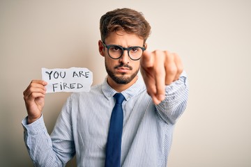 Young businessman wearing glasses holding paper with you are fired message pointing with finger to the camera and to you, hand sign, positive and confident gesture from the front