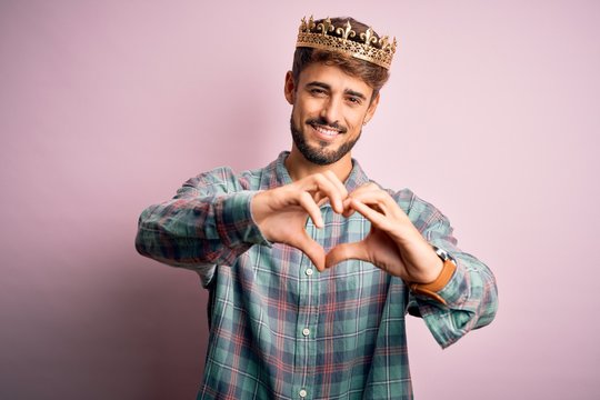 Young man with beard wearing golden crown of king standing over isolated pink background smiling in love showing heart symbol and shape with hands. Romantic concept.