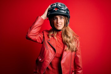 Young beautiful brunette motrocyclist woman wearing moto helmet over red background confuse and wonder about question. Uncertain with doubt, thinking with hand on head. Pensive concept.