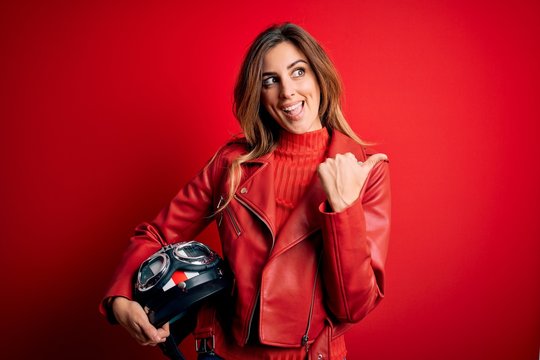 Beautiful brunette motorcyclist woman wearing red jacket holding motorcycle helmet pointing and showing with thumb up to the side with happy face smiling