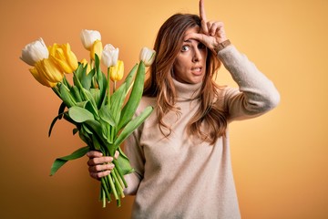 Young beautiful brunette woman holding bouquet of yellow tulips over isolated background making fun...