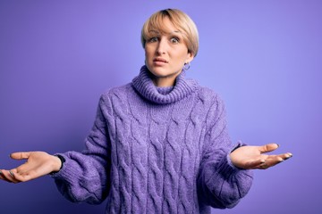 Young blonde woman with short hair wearing winter turtleneck sweater over purple background clueless and confused with open arms, no idea concept.