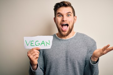 Young blond veggie man with beard and blue eyes holding paper with vegan message very happy and excited, winner expression celebrating victory screaming with big smile and raised hands