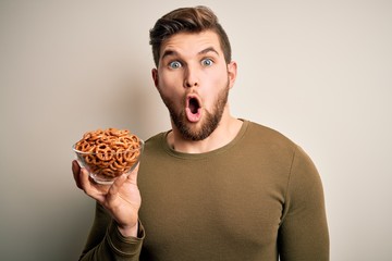 Young blond German man with beard and blue eyes holding bowl with baked pretzel scared in shock with a surprise face, afraid and excited with fear expression