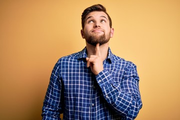 Young blond businessman with beard and blue eyes wearing shirt over yellow background Thinking concentrated about doubt with finger on chin and looking up wondering