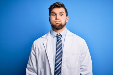 Young blond therapist man with beard and blue eyes wearing coat and tie over background puffing cheeks with funny face. Mouth inflated with air, crazy expression.