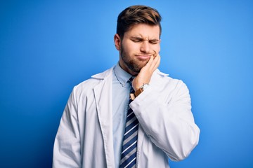 Young blond therapist man with beard and blue eyes wearing coat and tie over background touching mouth with hand with painful expression because of toothache or dental illness on teeth. Dentist