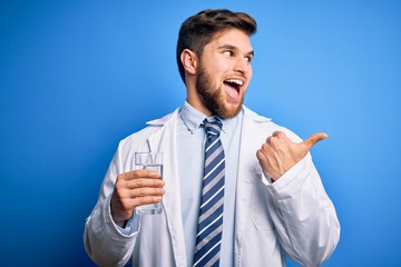 Young blond doctor man with beard and blue eyes wearing coat and tie drinking glass of water pointing and showing with thumb up to the side with happy face smiling