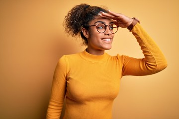 Fototapeta na wymiar Young beautiful african american girl wearing sweater and glasses over yellow background very happy and smiling looking far away with hand over head. Searching concept.