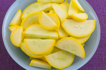 Sliced raw yellow summer crookneck squash in blue bowl on purple tablecloth