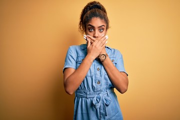 Young beautiful african american girl wearing denim dress standing over yellow background shocked covering mouth with hands for mistake. Secret concept.