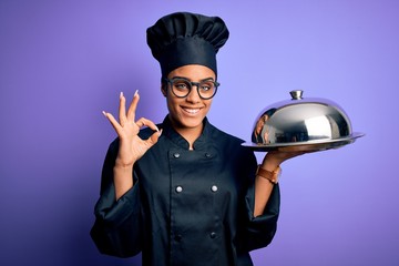 Young african american cooker girl wearing uniform and hat holding tray with dome doing ok sign with fingers, excellent symbol