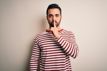 Young handsome man with beard wearing casual striped t-shirt standing over white background asking to be quiet with finger on lips. Silence and secret concept.