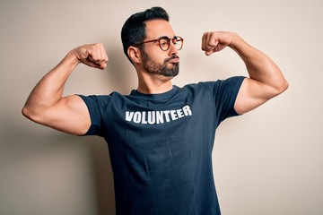 Handsome man with beard wearing t-shirt with volunteer message over white background showing arms muscles smiling proud. Fitness concept.