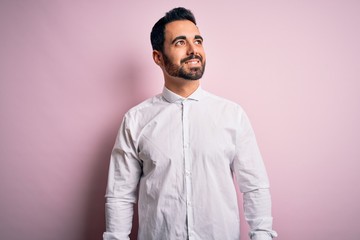 Young handsome man with beard wearing casual shirt standing over pink background looking away to side with smile on face, natural expression. Laughing confident.