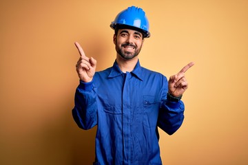 Mechanic man with beard wearing blue uniform and safety helmet over yellow background smiling confident pointing with fingers to different directions. Copy space for advertisement