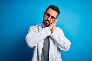 Young handsome doctor man with beard wearing coat and glasses over blue background sleeping tired dreaming and posing with hands together while smiling with closed eyes.