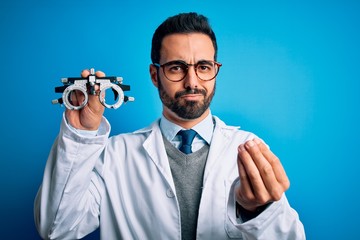Young handsome optical man with beard holding optometry glasses over blue background doing money gesture with hands, asking for salary payment, millionaire business