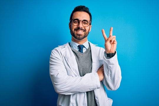 Young handsome doctor man with beard wearing coat and glasses over blue background smiling with happy face winking at the camera doing victory sign. Number two.
