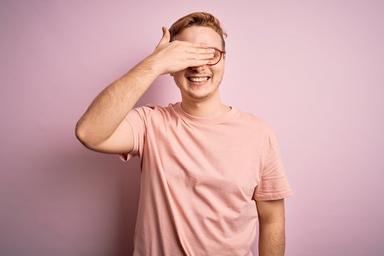 Young handsome redhead man wearing casual t-shirt standing over isolated pink background smiling and laughing with hand on face covering eyes for surprise. Blind concept.