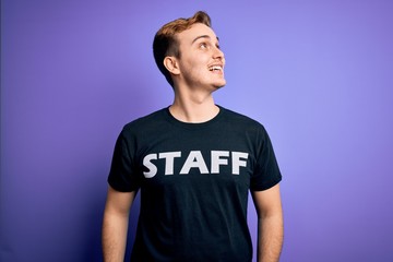 Young handsome redhead worker man wearing staff t-shirt uniform over purple background looking away to side with smile on face, natural expression. Laughing confident.