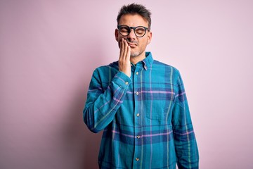 Young handsome man wearing casual shirt and glasses standing over isolated pink background touching mouth with hand with painful expression because of toothache or dental illness on teeth. Dentist