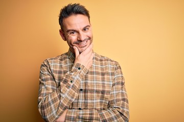 Young handsome man wearing casual shirt standing over isolated yellow background looking confident at the camera smiling with crossed arms and hand raised on chin. Thinking positive.