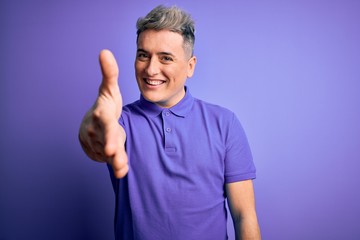 Young handsome modern man wearing casual purple t-shirt over isolated background smiling friendly offering handshake as greeting and welcoming. Successful business.
