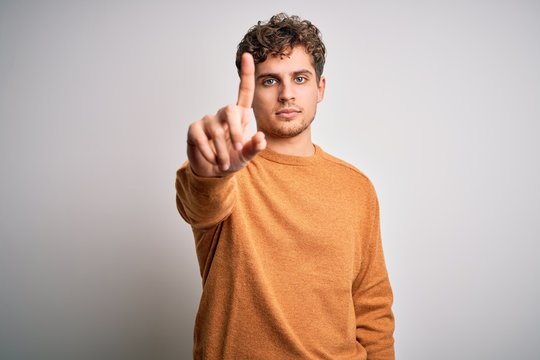 Young blond handsome man with curly hair wearing casual sweater over white background Pointing with finger up and angry expression, showing no gesture