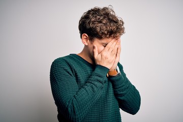 Young blond handsome man with curly hair wearing green sweater over white background with sad expression covering face with hands while crying. Depression concept.