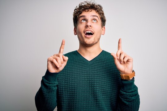 Young blond handsome man with curly hair wearing green sweater over white background amazed and surprised looking up and pointing with fingers and raised arms.
