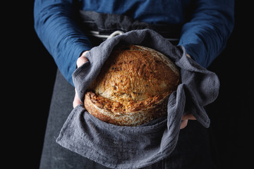 Baker holds in his hands in a towel fresh rustic bread with seeds, black background. Concept for...