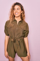 Young beautiful blonde woman with curly hair smiling happy and confident. Standing with smile on face wearing casual dress over isolated pink background