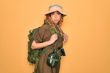 Young blonde explorer woman with blue eyes hiking wearing backpack and water canteen puffing cheeks with funny face. Mouth inflated with air, crazy expression.
