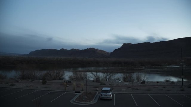 A timelapse looking out across a hotel car park and the Colorado River towards distant cliffs at sunset, as the light fades from the sky and the car park lights illuminate.