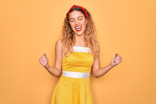 Beautiful blonde pin-up woman with blue eyes wearing diadem standing over yellow background very happy and excited doing winner gesture with arms raised, smiling and screaming for success. Celebration