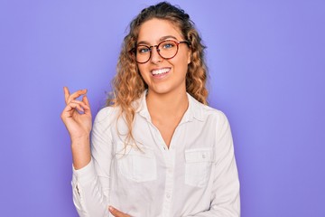 Young beautiful woman with blue eyes wearing casual shirt and glasses over purple background with a big smile on face, pointing with hand and finger to the side looking at the camera.