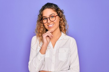 Young beautiful woman with blue eyes wearing casual shirt and glasses over purple background looking confident at the camera with smile with crossed arms and hand raised on chin. Thinking positive.