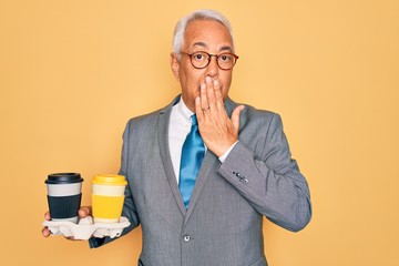 Middle age senior grey-haired business man wearing glasses holding coffee cup on tray cover mouth with hand shocked with shame for mistake, expression of fear, scared in silence, secret concept