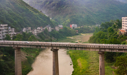 Naklejka premium Yichang, China - May 5, 2010: Bridge on pillars over brown meandering rivers between green mountain slopes with houses along a road.