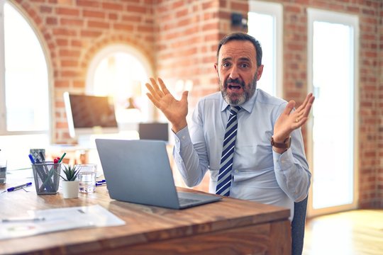 Middle age handsome businessman wearing tie sitting using laptop at the office clueless and confused expression with arms and hands raised. Doubt concept.