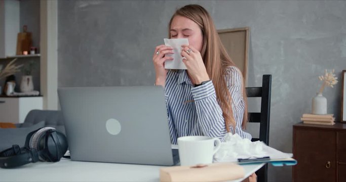 Beautiful young Caucasian blonde woman is sick with cold, sneezing while using laptop to work from home on quarantine.